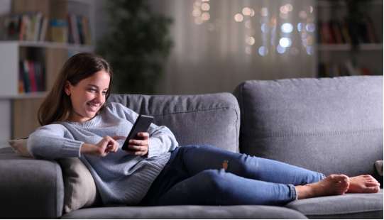 Mulher deitada em um sofa cinza segurando celular